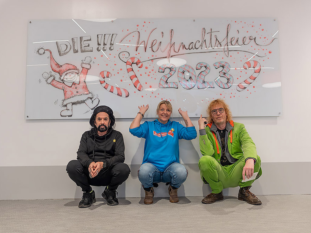 Christof Stein-Schneider, Susi Duhme als Organisatorin und Ecki Stieg von Fury in the Slaughterhouse vor dem Whiteboard mit der Aufschrift<br> „DIE!!! Weihnachtsfeier 2023”
